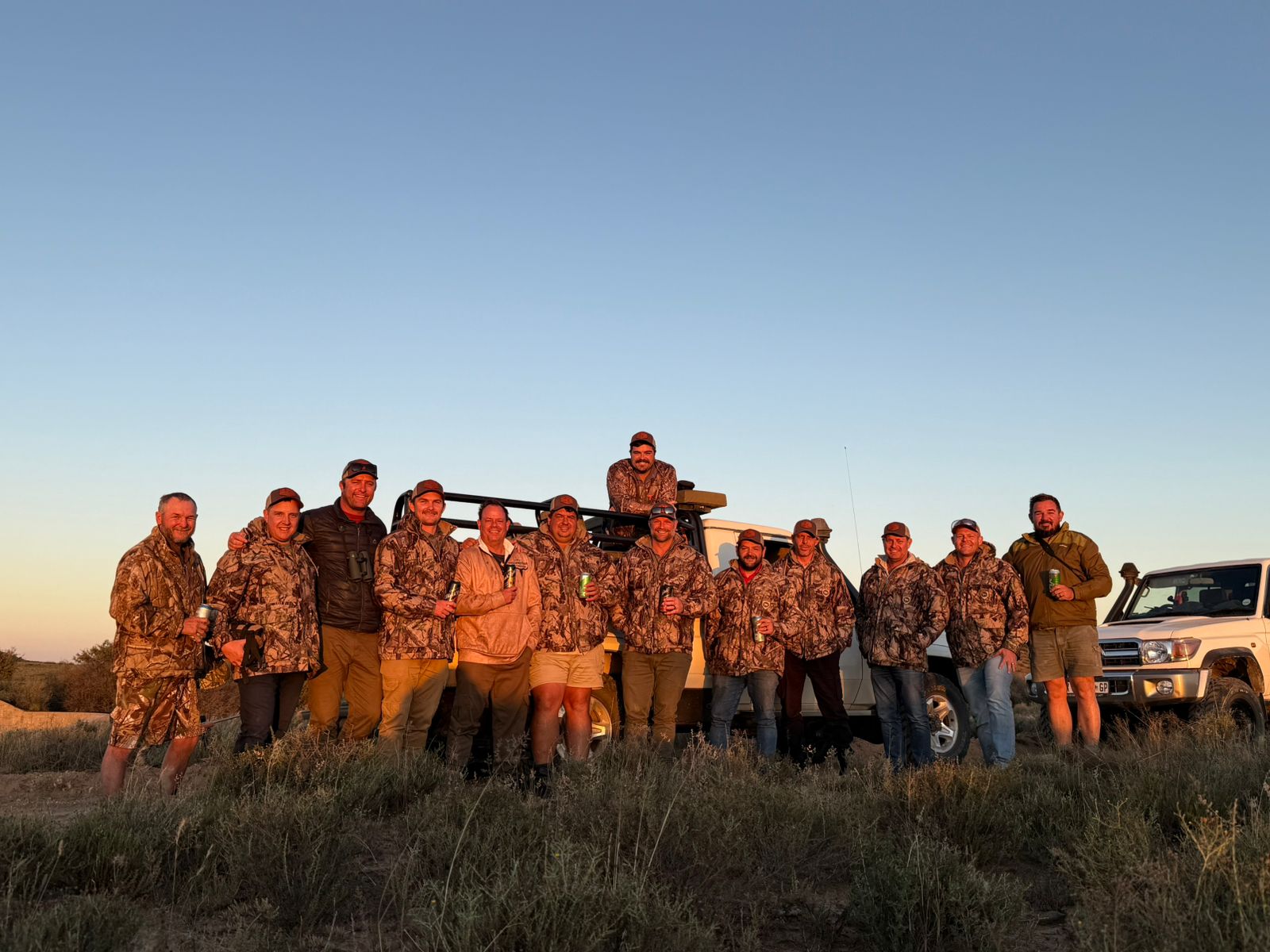 A group of fourteen in camouflage jackets and caps standing together in front of two vehicles on open veld at last light