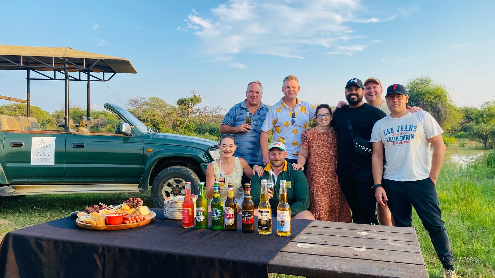 Eight guests standing together beside an open green game viewer at a sundowner table set with a grazing platter, a waterhole and bush behind them