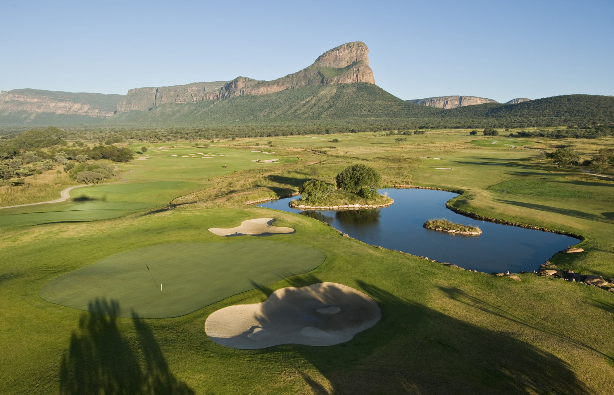An elevated view across a green, bunkers and a lake on a bushveld course, the Waterberg escarpment and a rock peak rising behind it