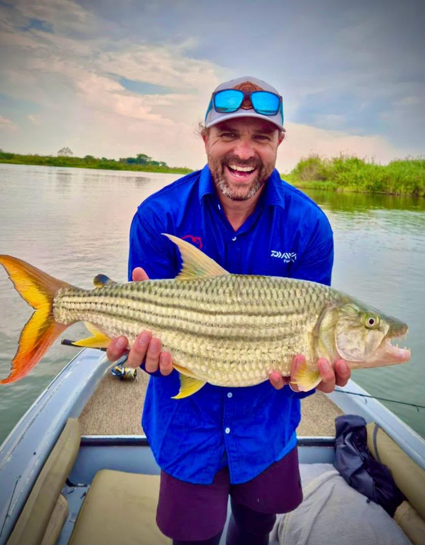 An angler in a boat holding a tiger fish, the river and a reed bank behind him