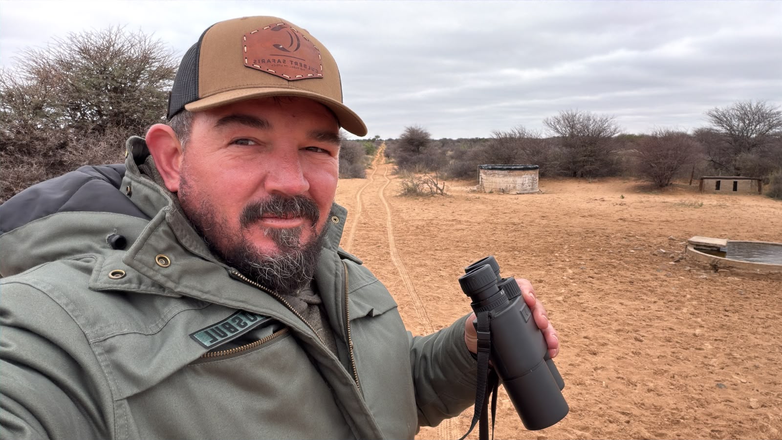 Chris Culbert in a field jacket and cap, binoculars in hand, on a sand track in the bushveld