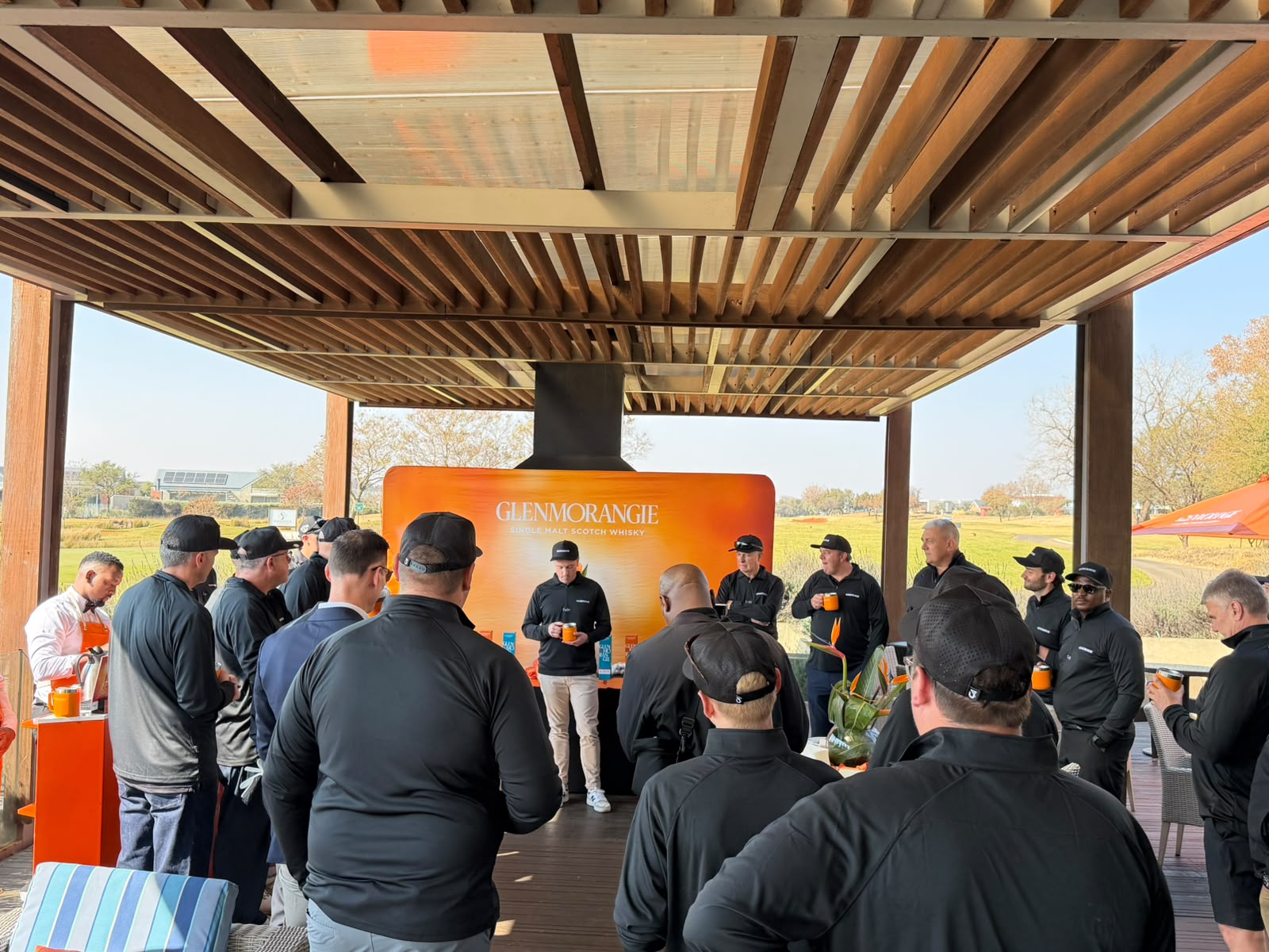 A corporate group gathered under a timber pergola at a golf course for a whisky tasting, presenter standing at a branded backdrop with the fairway behind